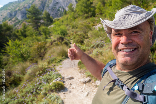 A middle-aged male hiker takes a selfie looking at the camera wearing a cap on a sunny day.