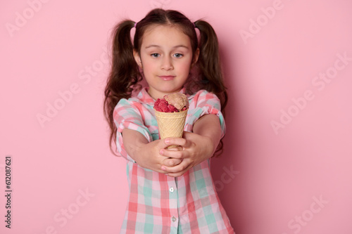 Caucasian beautiful smiling little kid girl with two ponytails, in checkered shirt, holding out at camera a waffle cone with delicious chocolate strawberry ice cream, isolated over pink background