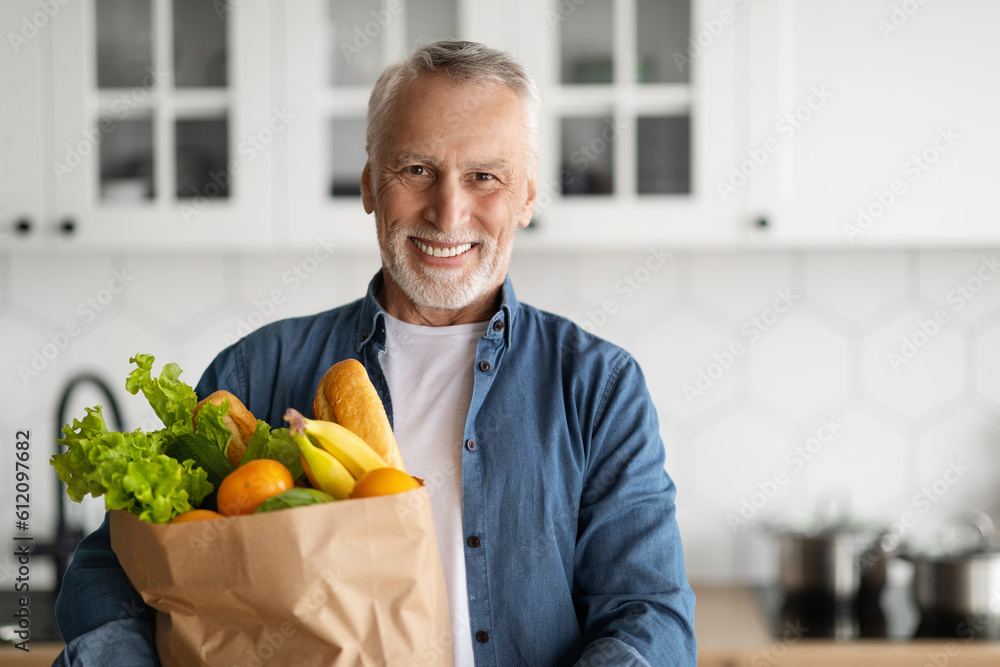 Fototapeta premium Happy Retirement. Smiling Senior Man Posing With Grocery Bag In Kitchen Interior