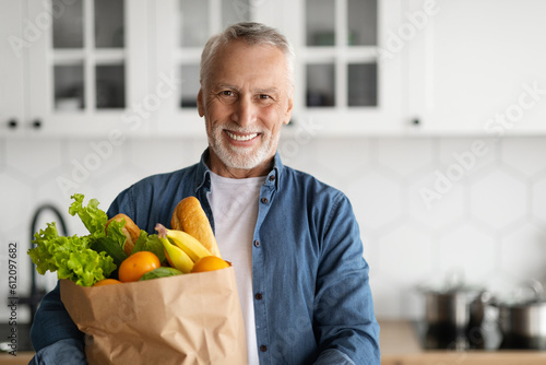 Happy Retirement. Smiling Senior Man Posing With Grocery Bag In Kitchen Interior