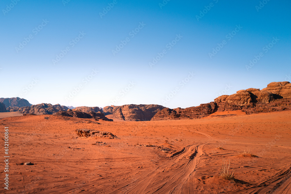 A sunny desert day in wadi rum national park, Jordan, with orange sand and tire tracks and a bright blue sky, a beautiful rocky landscape in the background and dunes in the foreground