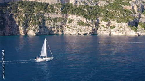 Aerial view of sailboat on blue sea at sunset in summer. Travel in Sardinia, Italy. Tropical seascape with sailing boat, sea bay, mountain, green trees, ocean, sky. Top drone view yacht. Yachting