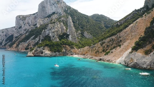 Aerial view of rocks and mountains, sea bay, beach, boats in summer day. Cala Goloritze, Sardinia Island, Italy. Top drone view of cliffs, stones, green trees, transparent azure water at sunset