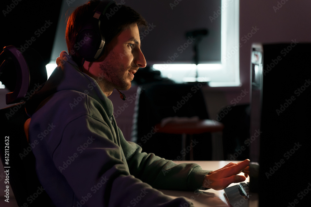 Side view of concentrated male eSport gamer in headphones with microphone sitting at white table and playing video game on desktop in dark room