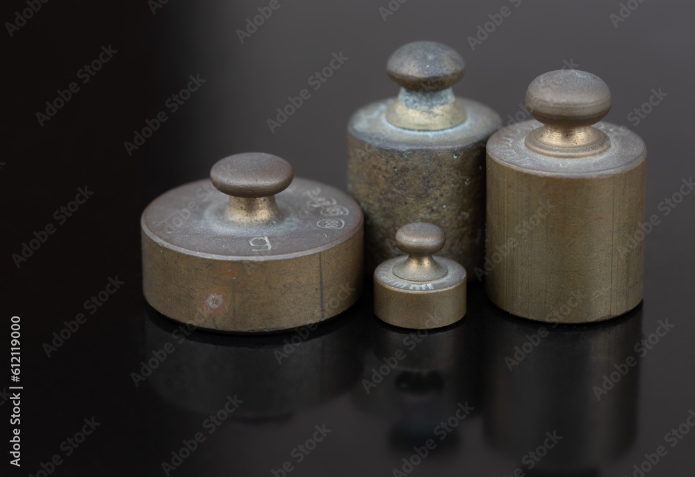 Four metal weights stand next to each other in front of a dark background and are reflected on the ground. These are small weights for a scale Four metal weights stand next to each other in front of a dark background and are reflected on the ground. These are small weights for a scale