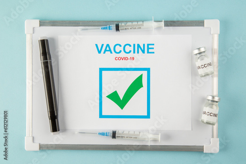 Above view of a sheet with VACCINE covid- tick and disposable syringes medicine ampoules on white desk on blue background photo