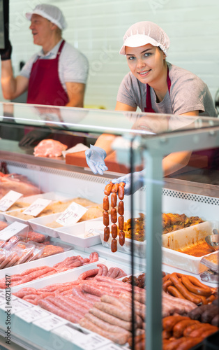 Young woman seller in uniform lays out meat sausages in butcher shop window