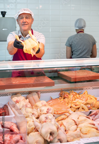 Adult man butcher in apron lays out raw chicken meat on counter in butcher shop