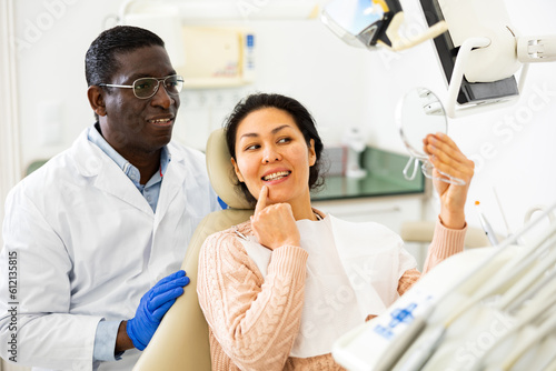 Positive asian woman and african-american man dentist looking at teeth through mirror after dental restoration procedure.