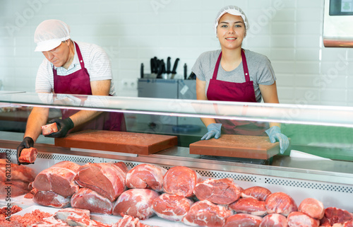 In organic natural meat store, friendly farmer female seller stands near counter refrigerator display case. In butcher shop, young female vendor in uniform apron, gloves, cap holds cutting board