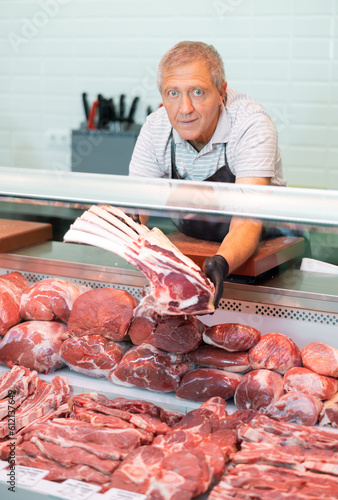 Positive elderly salesman in apron and disposable gloves shows raw beef tomahawk steak . Senior male farmer of meat products in butcher shop offers to buy veal flat ribs