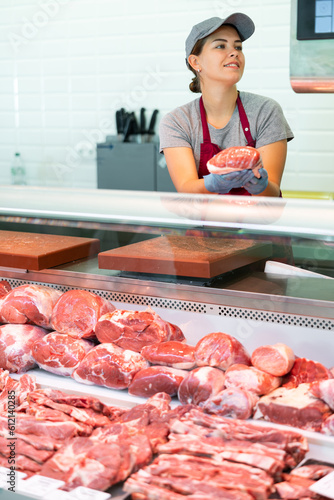 Positive young girl, professional butcher shop saleswoman, arranging meat products in display case, laying out fresh raw beef
