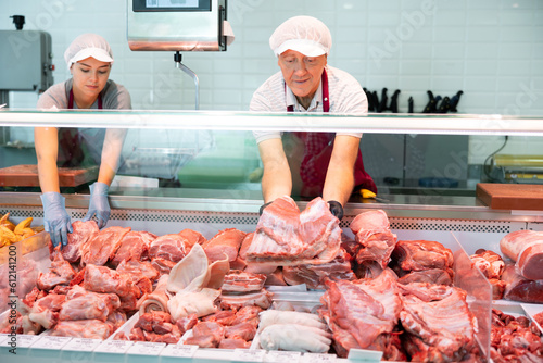 Positive skilled elderly butcher shop seller arranging meat products in glass refrigerated display case, laying out slab of fresh raw pork ribs