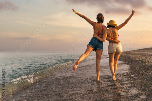 Sunset Beach Stroll: Joyful Friends Embracing Adventure - Two young friends, one wearing a straw hat, walk on the beach at sunset, raising their hands in joyful gestures.