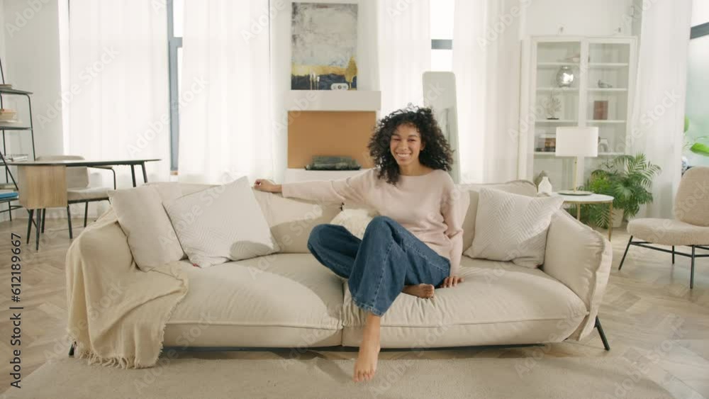 Happy African American woman jumping on modern soft sofa. Curly girl laughing, enjoying rest on cozy couch, feeling overjoyed, celebrating move in day to home, rented flat. Design interior furniture Happy African American woman jumping on modern soft sofa. Curly girl laughing, enjoying rest on cozy couch, feeling overjoyed, celebrating move in day to home, rented flat. Design interior furniture