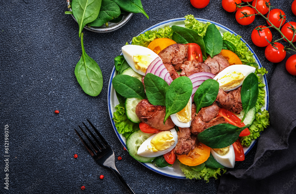 Perigord salad with roasted chicken liver, red tomatoes, cucumbers, boiled eggs, lettuce and spinach. Black kitchen table background, top view