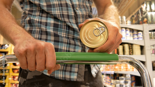 Close-up of a male buyer's hands with a shopping trolley holding a canned food