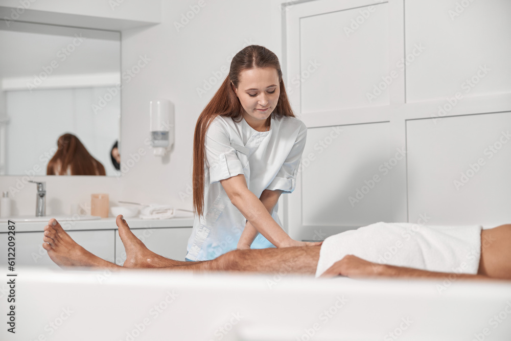 Smiling woman massages legs of mature man lying on couch in clinic Smiling woman massages legs of mature man lying on couch in clinic