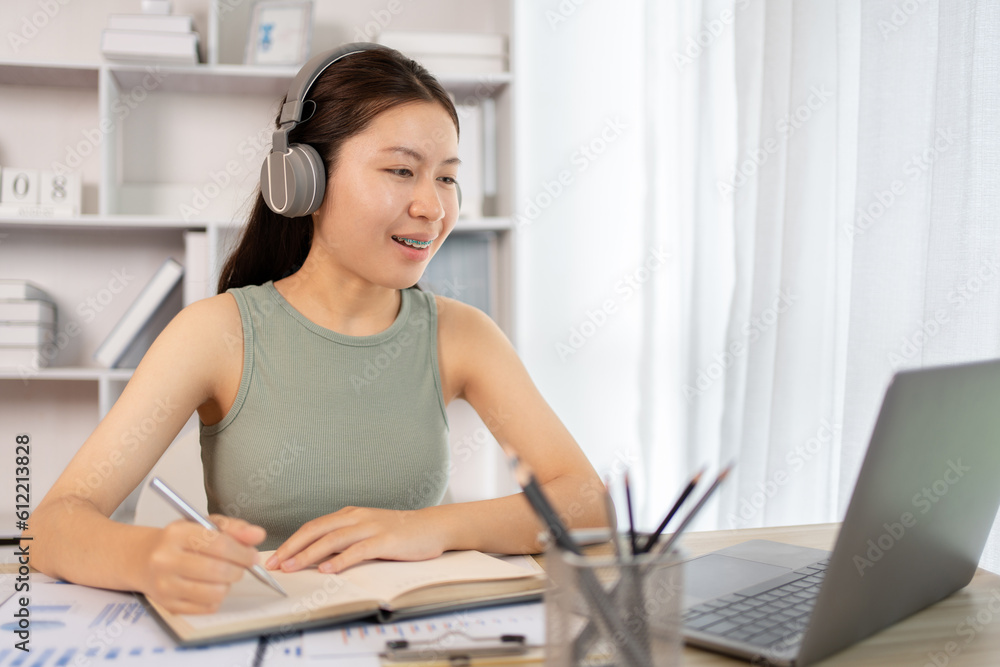 Beautiful young asian woman watching live video or video call of teacher teaching on laptop in her home, Take notes of important conversations and messages during the teacher's teaching. Beautiful young asian woman watching live video or video call of teacher teaching on laptop in her home, Take notes of important conversations and messages during the teacher's teaching.