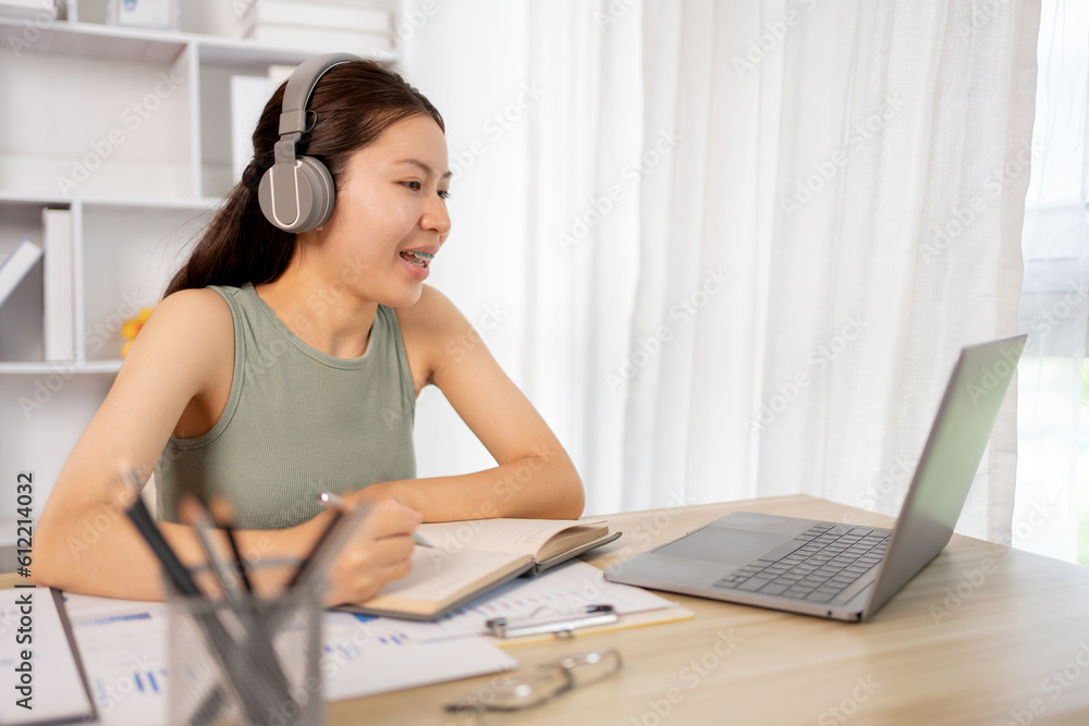 Obraz premium Beautiful young asian woman watching live video or video call of teacher teaching on laptop in her home, Take notes of important conversations and messages during the teacher's teaching.