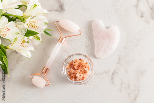 Quartz massage roller and gua sha scraper on a white marble background with astromeria flowers and a bowl of sea salt. Top view. A copy space. photo