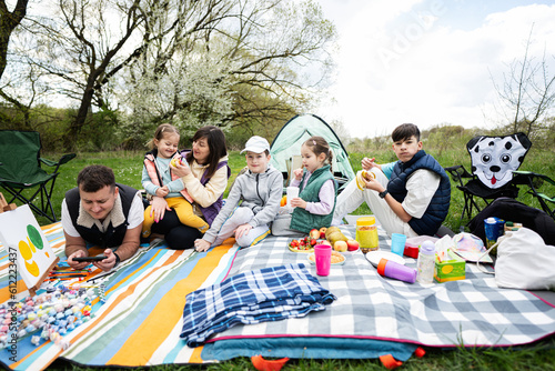 Happy young large family with four children having fun and enjoying outdoor on picnic blanket at garden spring park, relaxation.