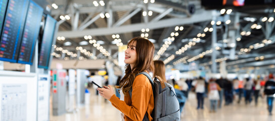 Young asian woman in international airport, using mobile smartphone and checking flight at the flight information board