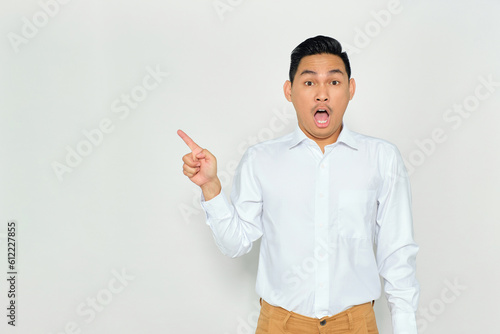 Portrait of surprised young Asian man in formal wear pointing fingers aside at copy space, presents something isolated on white background