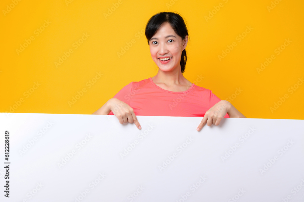 young asian sports fitness woman happy smile wearing pink sportswear standing behind the white blank banner or empty space advertisement board against yellow background. wellbeing lifestyle concept. young asian sports fitness woman happy smile wearing pink sportswear standing behind the white blank banner or empty space advertisement board against yellow background. wellbeing lifestyle concept.