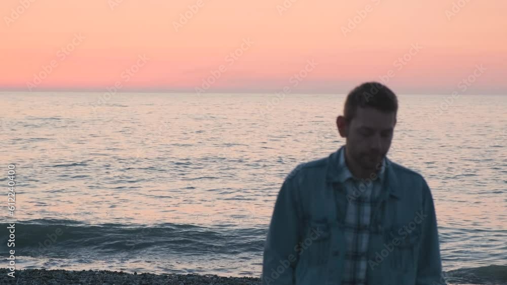 Young Caucasian man walking on the sea coast alone at sunset. Thoughtful pensive guy on the beach by the ocean. Shift focus. Loneliness, depression, melancholy