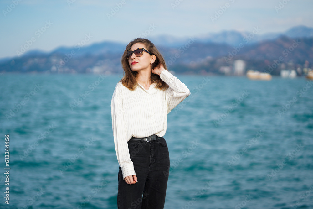 Sea Walk. Young woman wearing sunglasses and casual clothing, posing by sea on Seascape with mountains. Portrait of happy woman walking outdoor in sea. Girl with Shirt and jeans standing on embankmen. Sea Walk. Young woman wearing sunglasses and casual clothing, posing by sea on Seascape with mountains. Portrait of happy woman walking outdoor in sea. Girl with Shirt and jeans standing on embankmen.