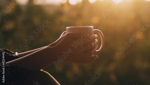 Close-up female hands holding a white ceramic cup of hot coffee or tea to drink at sunrise outdoors. Steam above the mug on blurred nature background. Peaceful morning concept.
