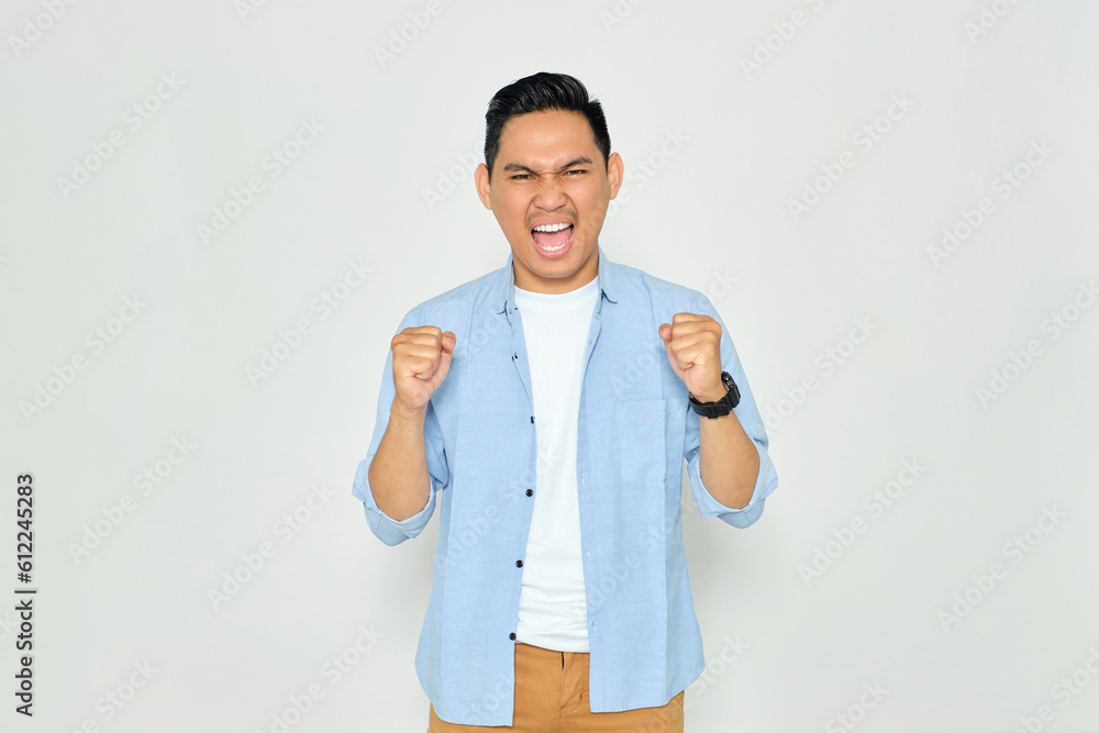 Excited young handsome Asian man in casual clothes making winner gesture with raising fist, celebrating success isolated on white background. Goal achievement concept Excited young handsome Asian man in casual clothes making winner gesture with raising fist, celebrating success isolated on white background. Goal achievement concept