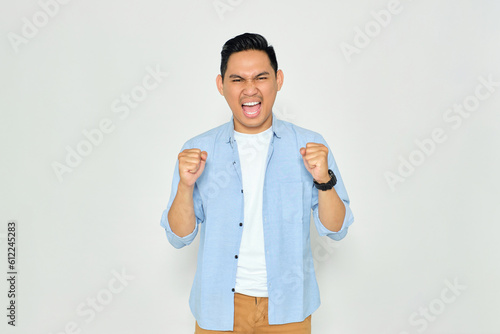 Excited young handsome Asian man in casual clothes making winner gesture with raising fist, celebrating success isolated on white background. Goal achievement concept