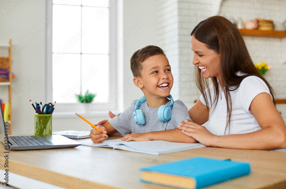 Joyful young mother at home cheerfully helps her first-grader son to do his homework. Woman and little boy are sitting at table with notebook and laptop and laughing looking at each other. Joyful young mother at home cheerfully helps her first-grader son to do his homework. Woman and little boy are sitting at table with notebook and laptop and laughing looking at each other.