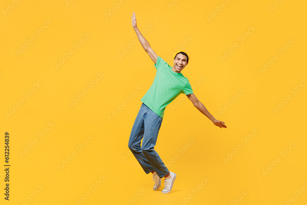 Full body young man of African American ethnicity he wear casual clothes green t-shirt hat stand on toes leaning back with outstretched hands dance isolated on plain yellow background studio portrait