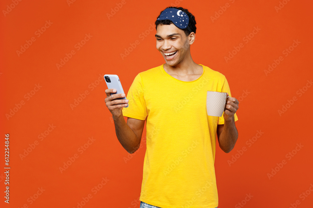 Calm young smiling happy man wear pyjamas jam sleep eye mask rest relax at home hold use mobile cell phone drink coffee isolated on plain orange background studio portrait Good mood night nap concept Calm young smiling happy man wear pyjamas jam sleep eye mask rest relax at home hold use mobile cell phone drink coffee isolated on plain orange background studio portrait Good mood night nap concept