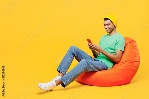 Full body fun young man of African American ethnicity he wears casual clothes green t-shirt hat sit in bag chair hold in hand use mobile cell phone isolated on plain yellow background studio portrait. © ViDi Studio