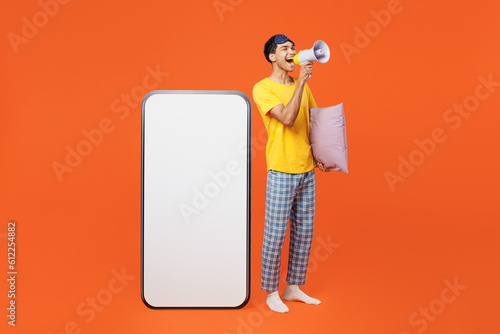 Full body young man in pyjamas jam sleep eye mask rest relax at home big huge blank screen area mobile cell phone smartphone scream in megaphone isolated on plain orange background Night nap concept