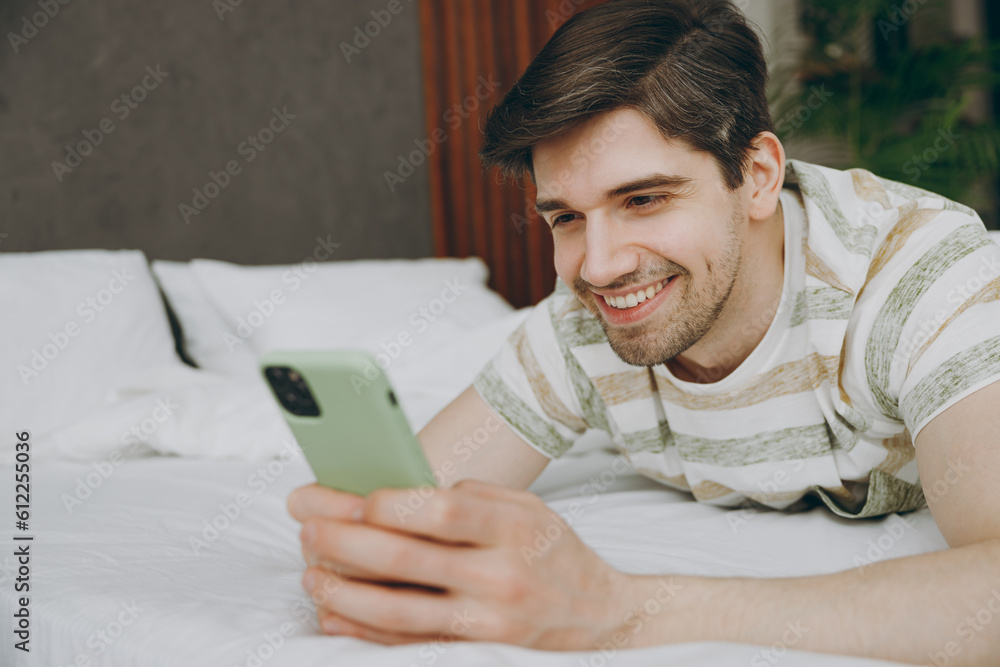 Young cheerful man wear casual clothes t-shirt pajama lying in bed hold in hand use mobile cell phone rest relax spend time in bedroom home in own room hotel wake up dream be in reverie good mood day. Young cheerful man wear casual clothes t-shirt pajama lying in bed hold in hand use mobile cell phone rest relax spend time in bedroom home in own room hotel wake up dream be in reverie good mood day.