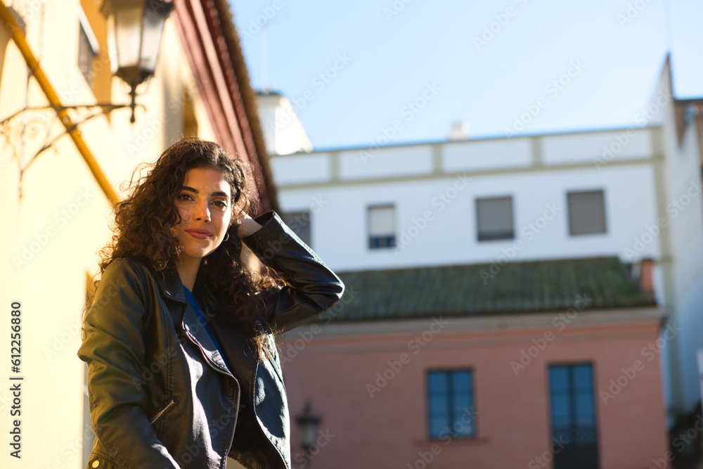 Young and beautiful Hispanic brunette woman with curly hair and black leather clothes is sitting on a railing in the street. The woman is happy and smiling.
