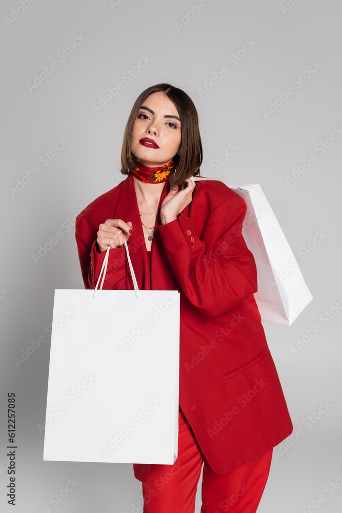consumerism, bold makeup, young woman with brunette short hair and nose piercing holding shopping bags and standing on grey background, youth culture, fashionable outfit, red suit