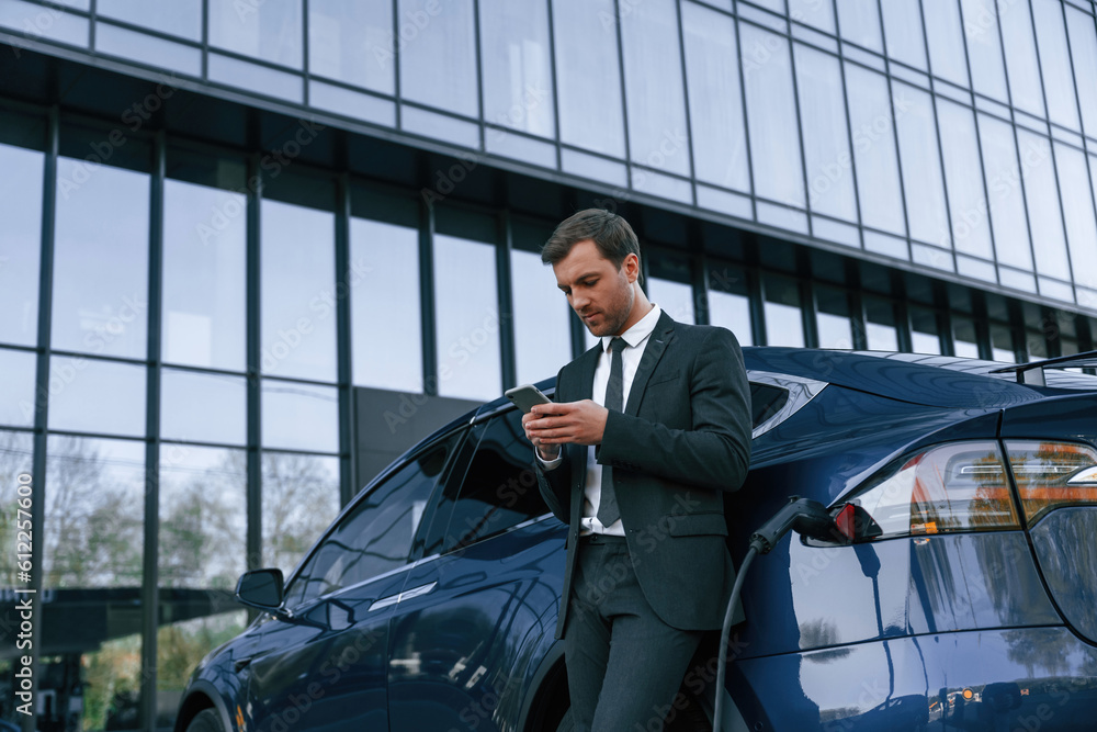 Using smartphone. Businessman is standing near his car outdoors
