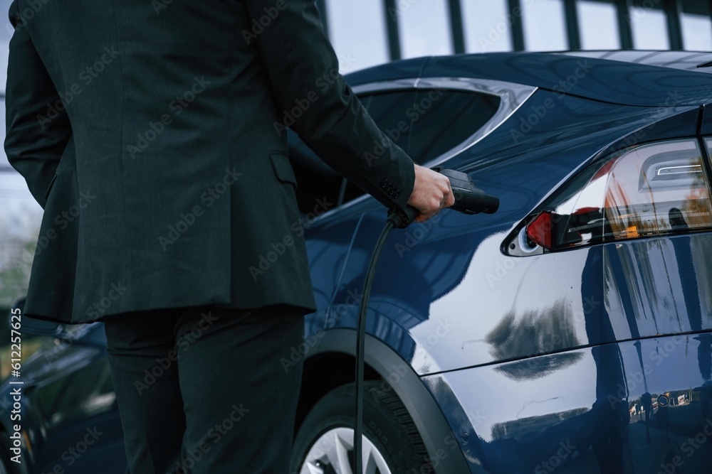 Close up view, putting charger into the socket. Businessman is standing near his electric car outdoors Close up view, putting charger into the socket. Businessman is standing near his electric car outdoors