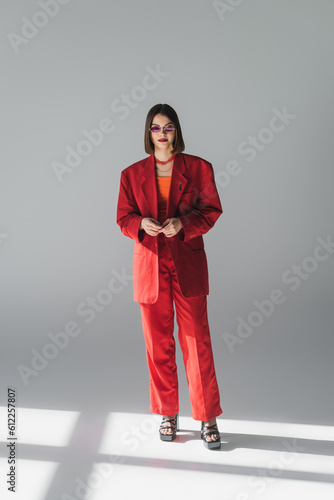 formal attire, young brunette woman with short hair posing in pink sunglasses and red suit on grey background, generation z, trendy outfit, fashionable model, full length