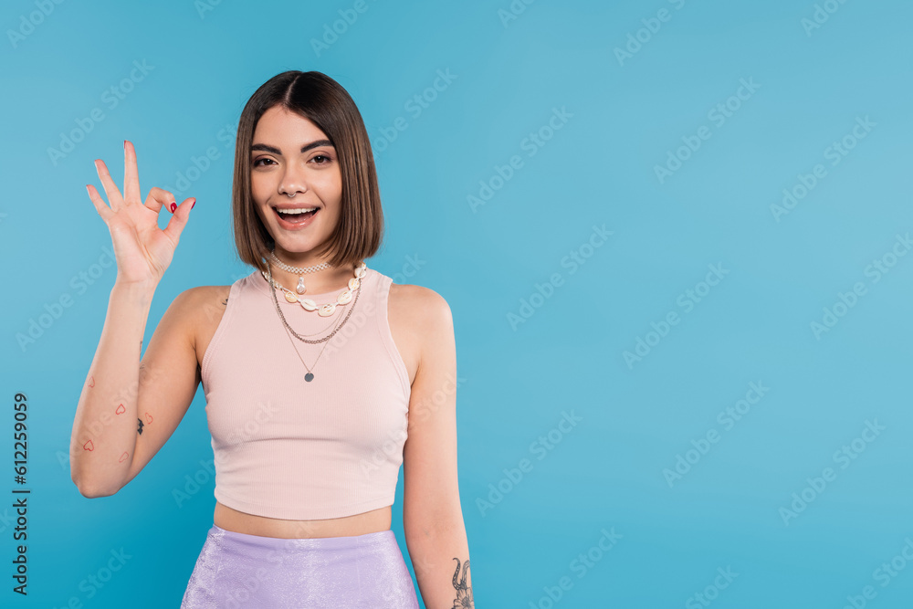 excitement, tattooed young woman with short hair in tank top smiling and showing ok gesture on blue background, casual attire, gen z fashion, fashionable trend, happiness excitement, tattooed young woman with short hair in tank top smiling and showing ok gesture on blue background, casual attire, gen z fashion, fashionable trend, happiness