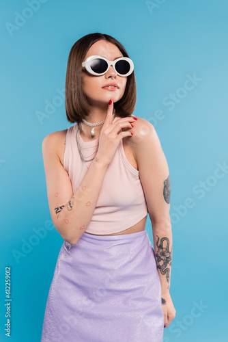 fashion trend, brunette young woman with short hair in tank top, skirt and sunglasses posing on blue background, casual attire, gen z fashion, personal style, everyday makeup, casual attire © LIGHTFIELD STUDIOS