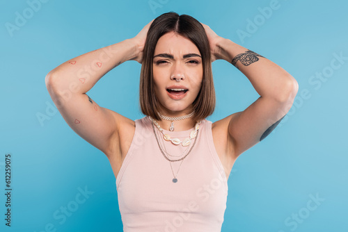 failure, upset young woman with short brunette hair and tattoos holding hands near head and looking at camera on blue background, casual attire, gen z fashion, emotional