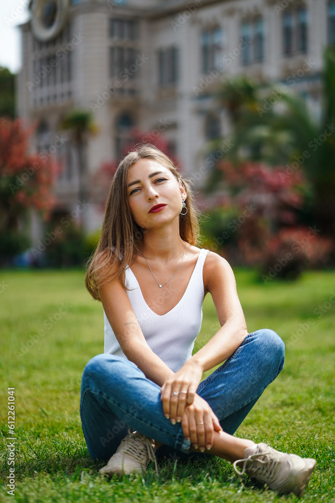 Beautiful young woman wearing tank top and jeans sitting on grass lawn in front of building. Young woman posing sitting on green grass. City recreation. Carefree person resting experiencing calmness
