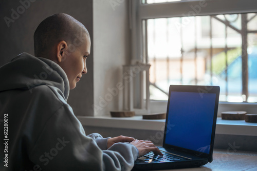 a beautiful young woman with a shaved bald head sits near the window and works in a laptop, communicates in social networks, works remotely